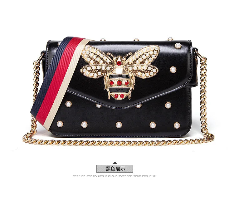 Black flap handbag with rhinestone bee clasp, pearl studs, striped strap, and gold chain.