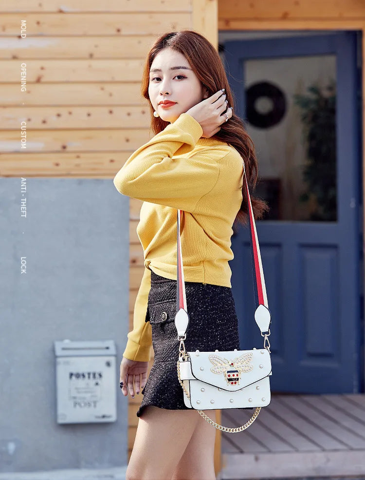 White crossbody bag with striped webbing strap and gold chain, with bee bow flap